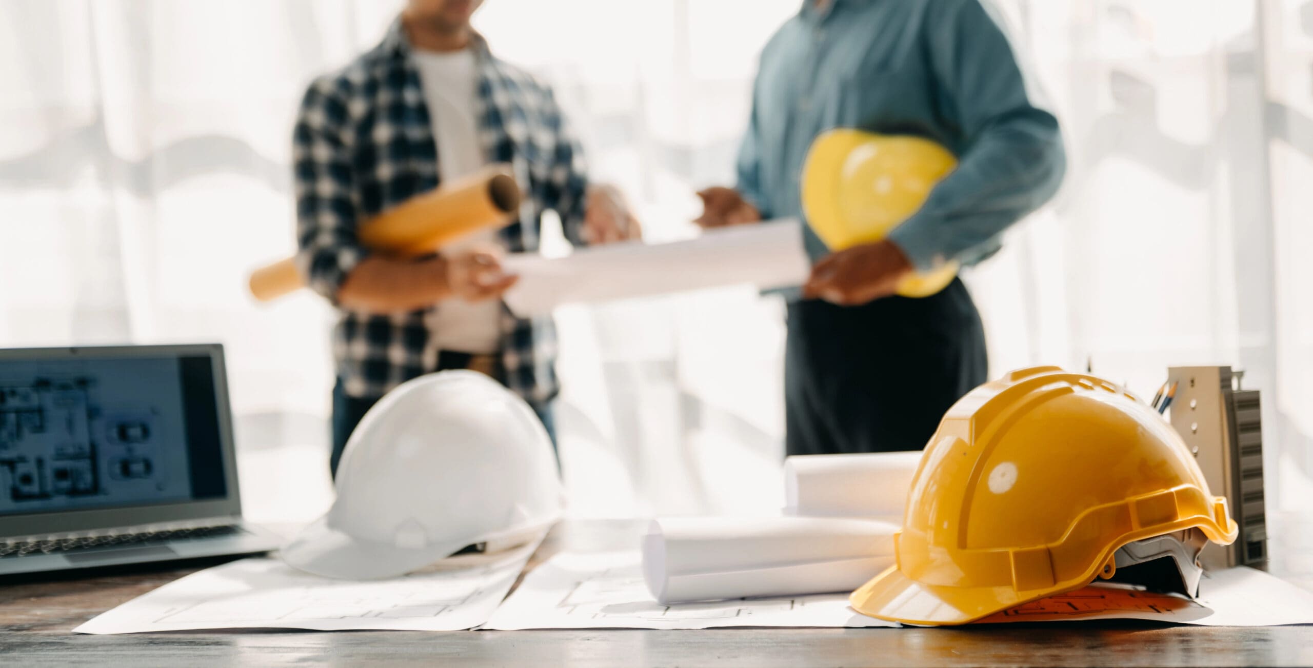 Two people in discussion holding blueprints. Hard hats and rolled plans on a table.