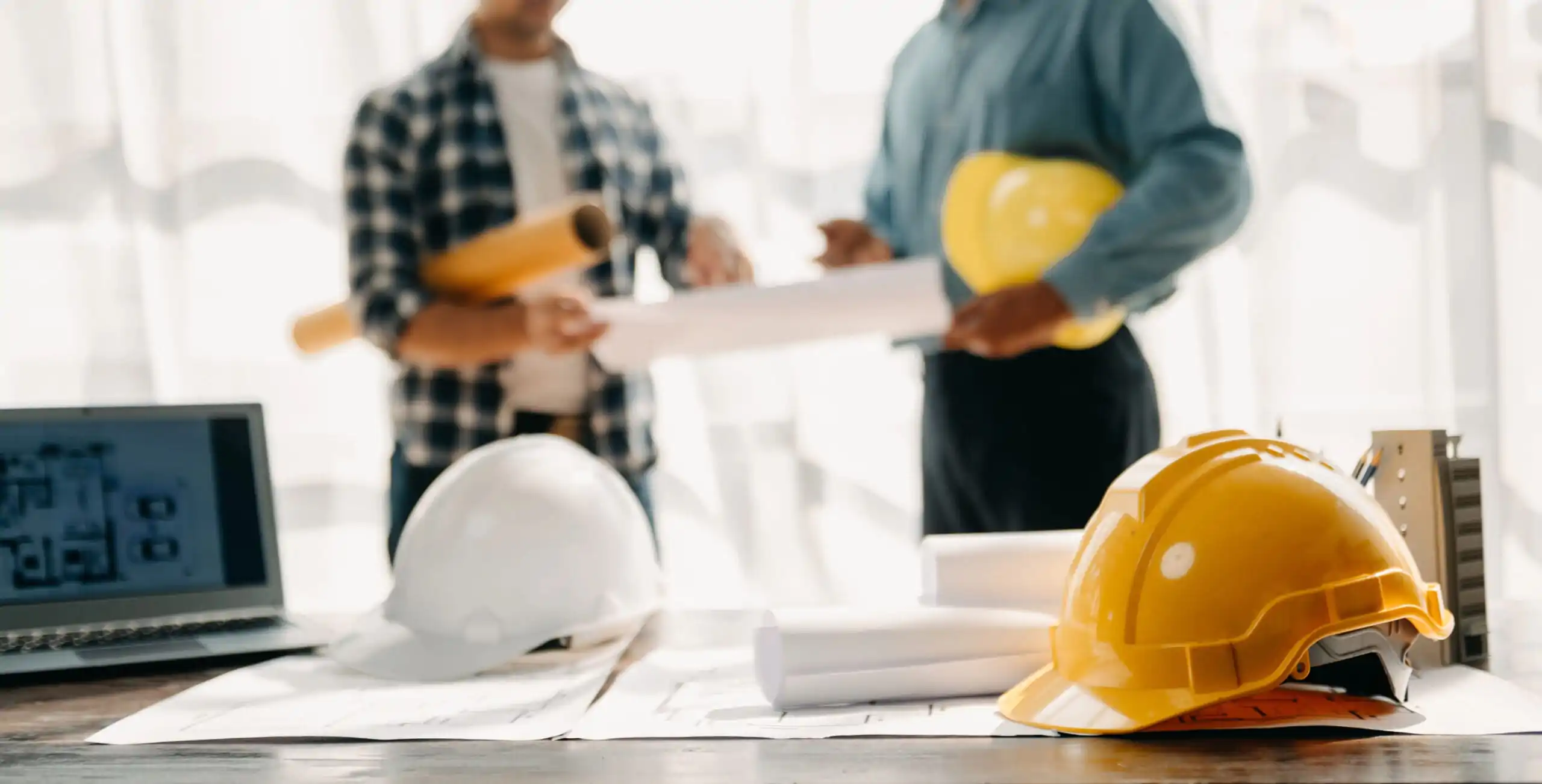 Two people in discussion holding blueprints. Hard hats and rolled plans on a table.