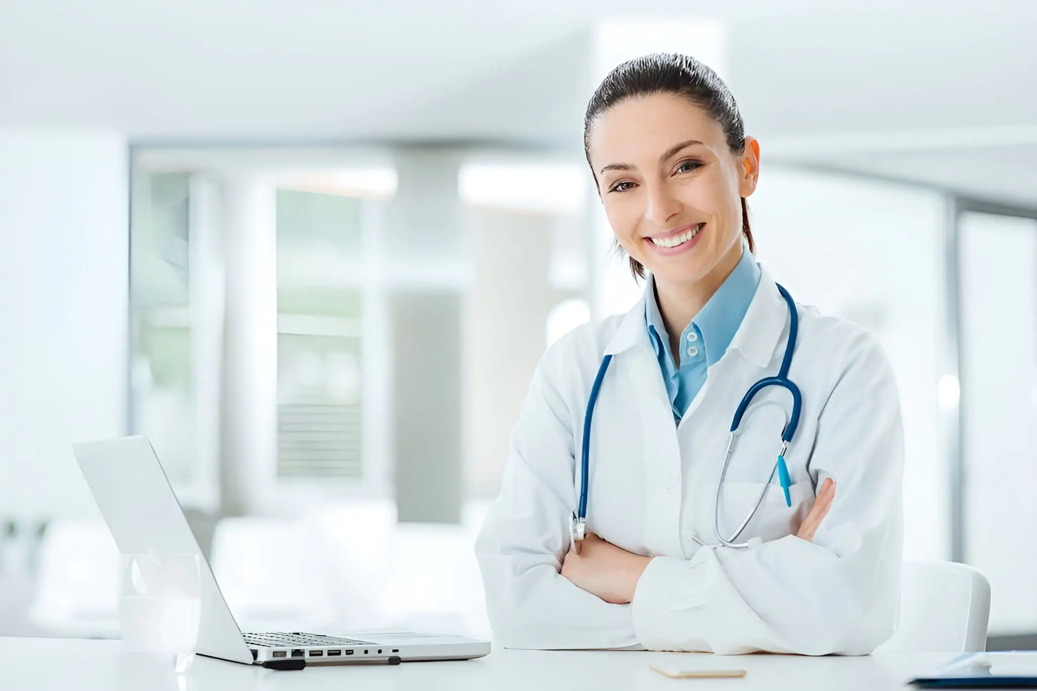 Smiling female doctor in a white coat with a stethoscope, sitting at a desk with a laptop.