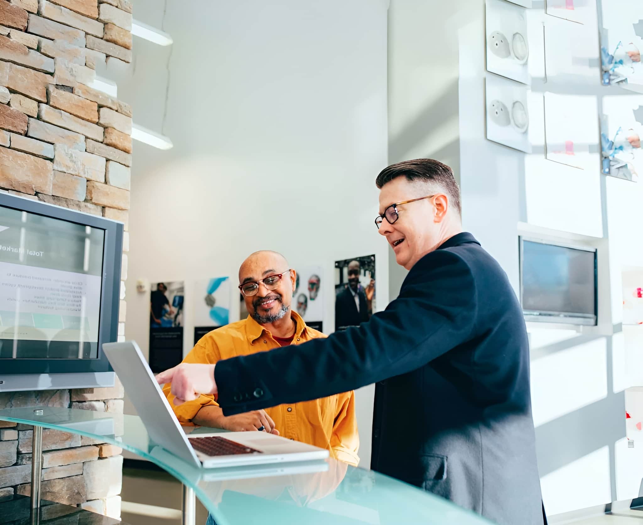 Two people in a modern office, one in a dark suit pointing at a laptop screen, and the other in a yellow shirt smiling.