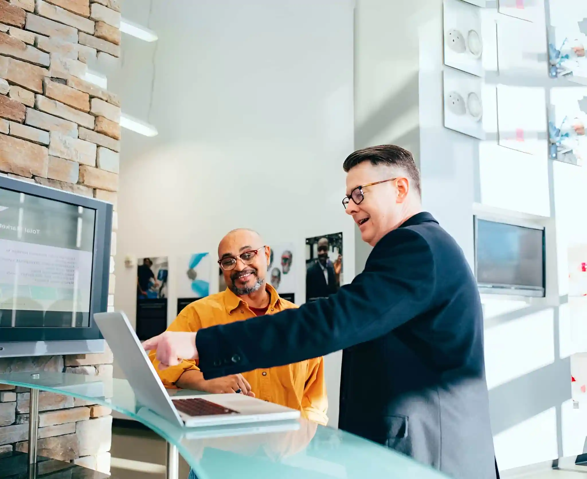 Two people in a modern office, one in a dark suit pointing at a laptop screen, and the other in a yellow shirt smiling.